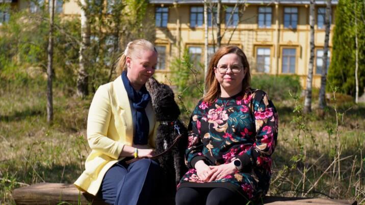 Kuvassa on kaksi naista ja musta koira istumassa puunrunkoon tehdyllä penkillä ulkona. Taustalla näkyy keltainen rakennus ja puita.