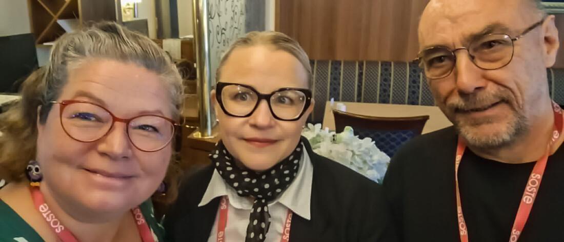 Kolme hymyilevää henkilöä yhteiskuvassa sisätiloissa. Heillä kaikilla on kaulassaan punaiset SOSTE-kaulanauhat, ja keskellä olevalla henkilöllä on lisäksi pinkki SOSTE-rintamerkki.