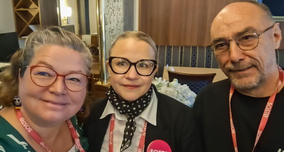 Kolme hymyilevää henkilöä yhteiskuvassa sisätiloissa. Heillä kaikilla on kaulassaan punaiset SOSTE-kaulanauhat, ja keskellä olevalla henkilöllä on lisäksi pinkki SOSTE-rintamerkki.