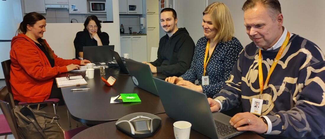 Viisi ihmistä istuu kokouspöydän ääressä työskentelemässä kannettavilla tietokoneilla. Pöydällä on tietokoneiden lisäksi kahvikuppeja, esitteitä ja kokousmikrofoni. Taustalla näkyy keittiötila.