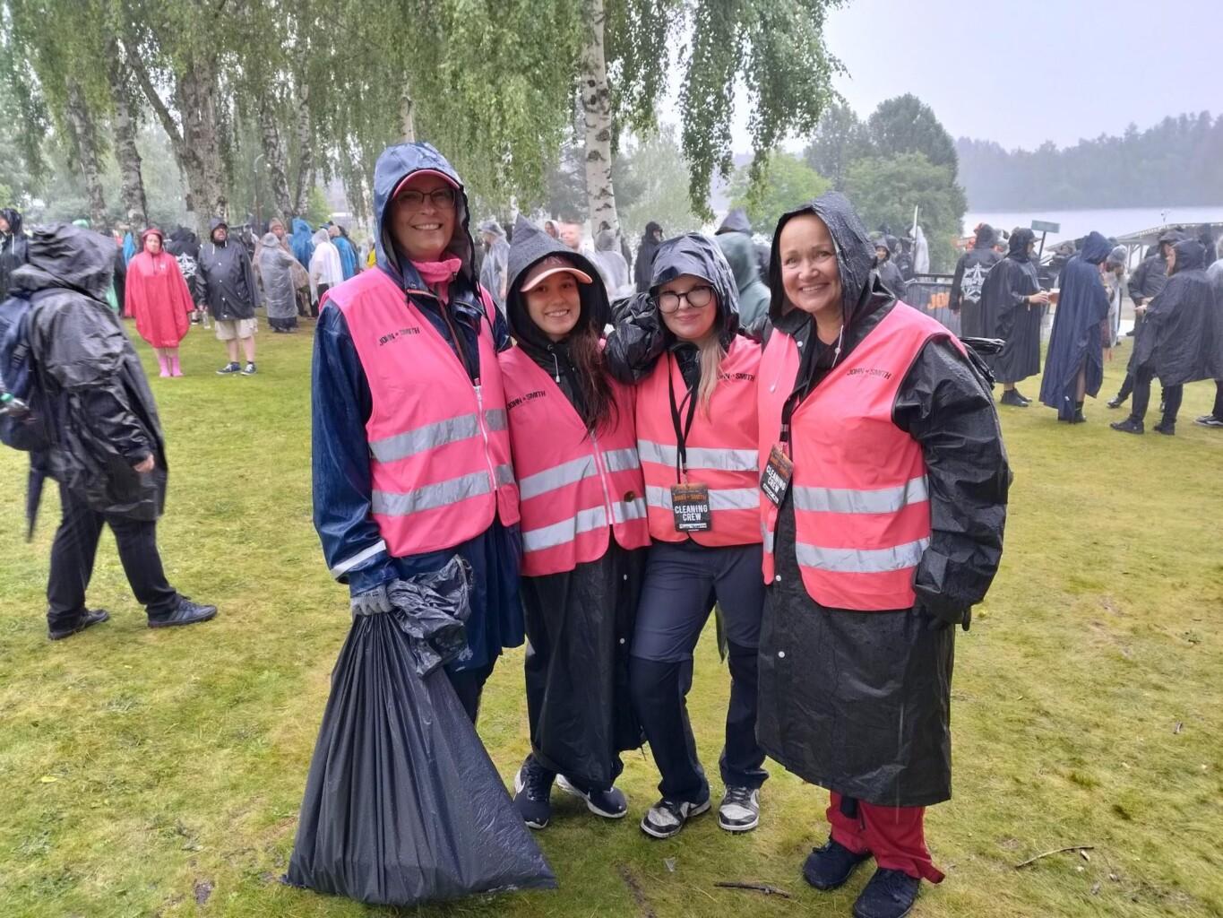Neljän hengen ryhmä vaaleanpunaisiin heijastinliiveihin ja tummiin sadevaatteisiin pukeutuneita henkilöitä hymyilee kameralle nurmikolla festivaalilla.